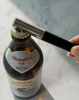 A close-up of a bottle opener held with a hand, with a black handle and silver metal blade, positioned to open a dark glass beer bottle with a white and gold cap. The opener is branded 'blomus'.