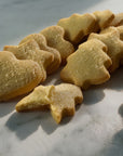A close-up shot of several golden shortbread-style butter cookies shaped like hearts, stars, and Christmas trees, dusted with crystallised sugar, arranged on a light marble surface under warm sunlight.