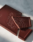 A dark chocolate bar with embossed decorative artwork and the brand name 'Coppeneur' visible in the upper left corner, photographed on a light marble surface with strong natural shadows.