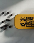 A yellow metal tin of Lemon and Ginger Liquorice sits on a light marble surface with several dark liquorice pearls scattered to the left. The tin features a black graphic of a bearded man wearing a beanie and the text 'REINES LAKRITS INGWER LIMONE'.