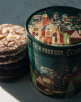 A stack of round gingerbread sits beside a decorated cylindrical tin labeled 'Nürnberger Elisen-Lebkuchen', photographed on a light marble surface with strong sunlight and shadows.
