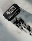 A small black metal tin labeled 'REINES LAKRITS' with a white logo, shown on a light marble surface with several black round liquorice beads spilled out beside it.