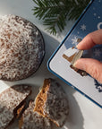 A hand is turning the silver dial of a blue music box-tin with a Christmas tree design. On a white surface next to the tin are decorated Christmas cookies, a stack whole and one broken to show the interior.