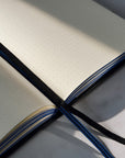 An open dotted journal with cream-colored pages, a black elastic closure, and a blue-and-black striped ribbon bookmark, photographed on a light surface with strong natural shadows.