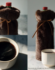 A dark brown faux-fur bottle cover shown wrapped around a wine bottle, with one bottle displayed upright and the other shown inside the cover. A small ceramic cup of mulled wine is also present on a light marble surface.