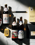 A styled flat-lay of beer bottles and accessories arranged on a white marble surface with dramatic diagonal shadows. Visible items include four dark brown bottles with labels, a black blomus bottle opener, a slab of chocolate in a black envelope, a small yellow Liquorice tin, and a yellow cookie box.