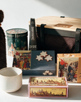 A curated gift basket arrangement on a white surface featuring a brown faux-fur bottle cover tied with a brown cord, a ceramic mug, a round tin, a dark hot chocolate stick, three boxes of cookies, and a large black mesh storage basket with black straps and wooden lid.