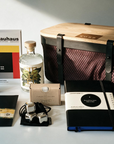 A curated collection of Bauhaus-inspired minimalist products arranged on a light surface, including a large black mesh basket with a natural wood lid, a Bauhaus book, a glass alcohol-free spirit bottle, a black notebook, a slab of chocolate in a black envelope, a small liquorice tin, and a small black pouch with metal ice cubes.