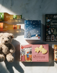 A styled flat-lay of assorted Christmas-themed treats and drinks on a light marble surface with strong sunlight and shadows. Items include two bottles of soda, a box of German Christmas butter-cookies, a music box-tin of gingerbread, a teddy bear, a hot-chocolate stick, a book of German Christmas stories, and fairytale house-decorated gingerbread box.
