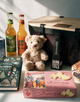 A curated gift basket arrangement featuring a light wood and black mesh basket, a teddy bear, two soda bottles, a music box-tin, a Christmas story book, a cookie box, a fairytale house-shaped box, and a hot chocolate stick, on a sunlit white surface.