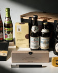 A curated beer gift set arranged on a dark surface with dramatic side lighting. Visible items include six beer bottles, a wooden-lidded storage case, a black blomus bottle opener with gift box, a slab of chocolate in a black envelope, a small yellow Liquorice tin, and a yellow cookie box.