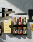 A styled flat-lay of beer bottles and accessories arranged on a white marble surface with dramatic diagonal shadows. Visible items include two dark brown bottles with labels, a clear bottle with a white label, three smaller bottles with colourful labels, a black blomus bottle opener, a slab of chocolate in a black envelope, a small yellow Liquorice tin, and a yellow cookie box.