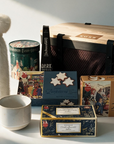A curated gift basket arrangement on a white surface featuring a white faux-fur bottle cover tied with a brown cord, a ceramic mug, a round tin, a dark hot chocolate stick, a box of stollen, two boxes of cookies, and a large black mesh storage basket with black straps and wooden lid.