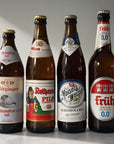 Four brown glass beer bottles with white labels, standing side by side on a light marble surface against a neutral background. The labels show different German beer brands and styles, including Flötzinger, Rothaus Pils, Maisel Weisse, and Früh Kölsch.