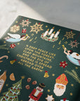 The rear of a dark green hardcover book with festive Christmas illustrations and a poem printed on the back in gold foil, photographed on a light marble surface with soft diagonal shadows.