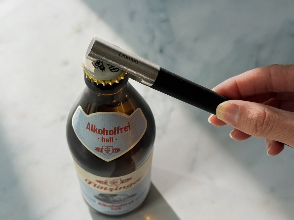 A close-up of a bottle opener held with a hand, with a black handle and silver metal blade, positioned to open a dark glass beer bottle with a white and gold cap. The opener is branded 'blomus'.