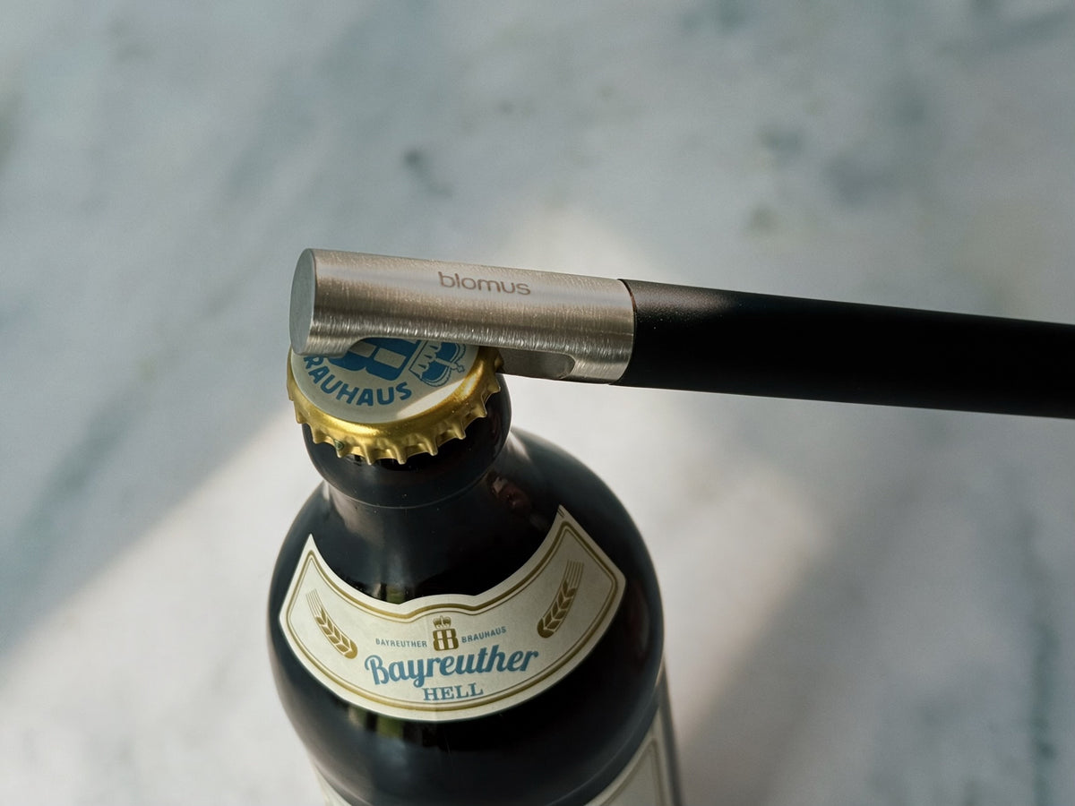 A close-up of a bottle opener with a black handle and silver metal blade, positioned to open a dark glass beer bottle with a gold cap. The opener is branded 'blomus'.