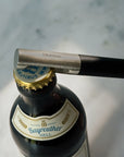 A close-up of a bottle opener with a black handle and silver metal blade, positioned to open a dark glass beer bottle with a gold cap. The opener is branded 'blomus'.