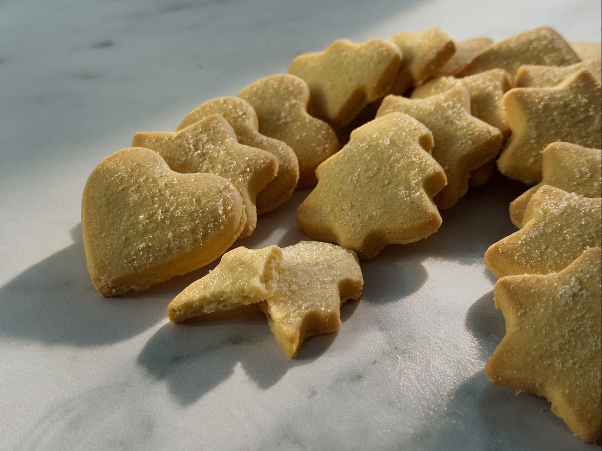 A close-up shot of several golden shortbread-style butter cookies shaped like hearts, stars, and Christmas trees, dusted with crystallised sugar, arranged on a light marble surface under warm sunlight.