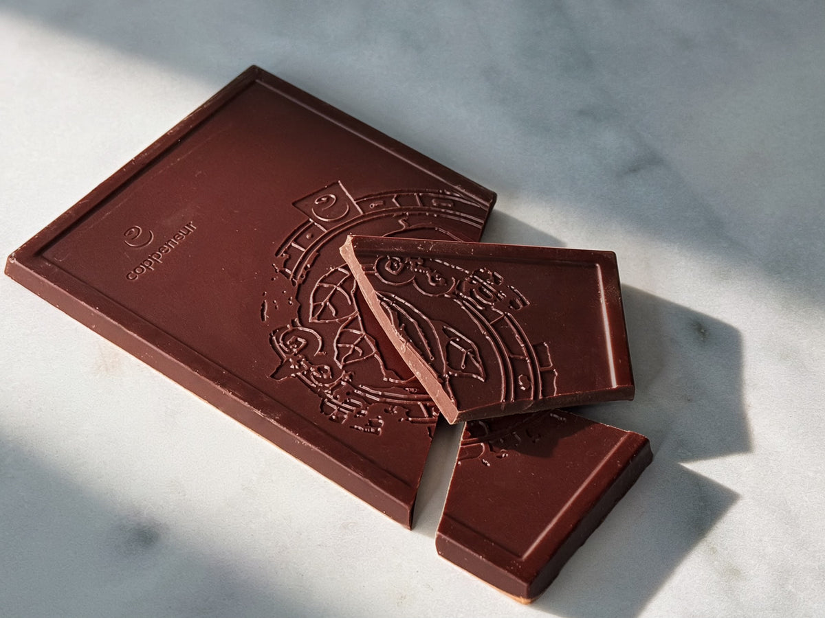 A dark chocolate bar with embossed decorative artwork and the brand name 'Coppeneur' visible in the upper left corner, photographed on a light marble surface with strong natural shadows.