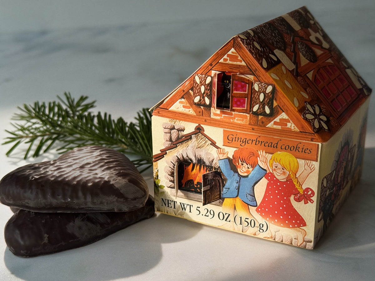 A Hansel &amp; Gretel inspired, house-shaped gingerbread cookie box with an illustrated cottage design, shown beside two dark chocolate gingerbread cookies on a light marble surface with a pine branch.