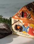 A Hansel & Gretel inspired, house-shaped gingerbread cookie box with an illustrated cottage design, shown beside two dark chocolate gingerbread cookies on a light marble surface with a pine branch.