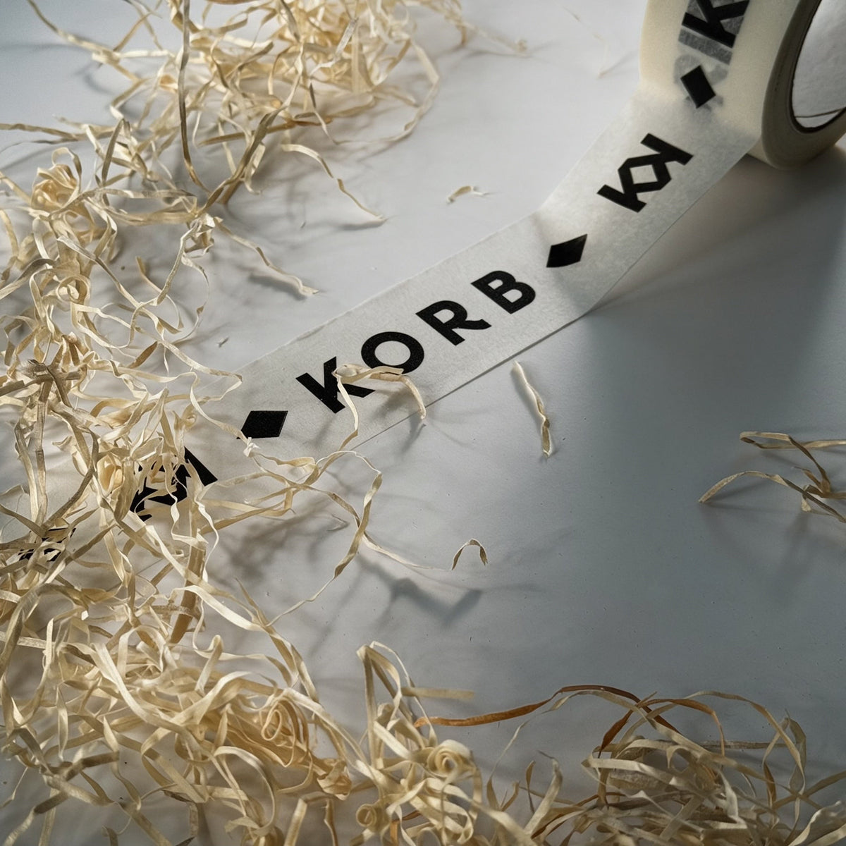 White Korb brand tape with black lettering and diamond symbols, partially unrolled over light wood shavings on a gray surface.