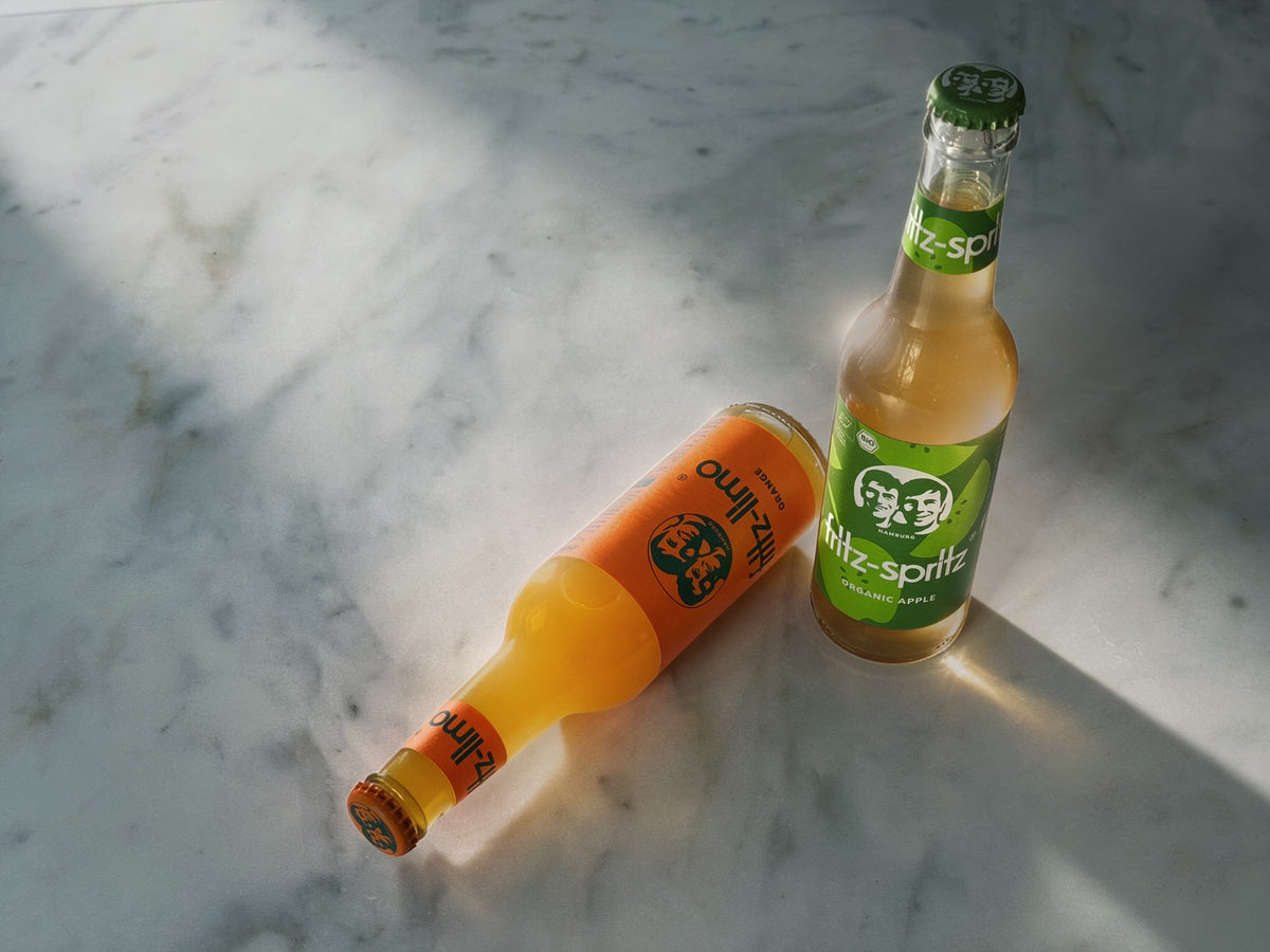 Two glass bottles of apple and orange soda on a white marble surface with strong sunlight and shadows. One bottle lies on its side filled with orange soda labelled Fritz-Limo Orange, and the other stands upright with pale apple juice and a green label reading 'Fritz-Spritz'.