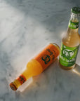 Two glass bottles of apple and orange soda on a white marble surface with strong sunlight and shadows. One bottle lies on its side filled with orange soda labelled Fritz-Limo Orange, and the other stands upright with pale apple juice and a green label reading 'Fritz-Spritz'.