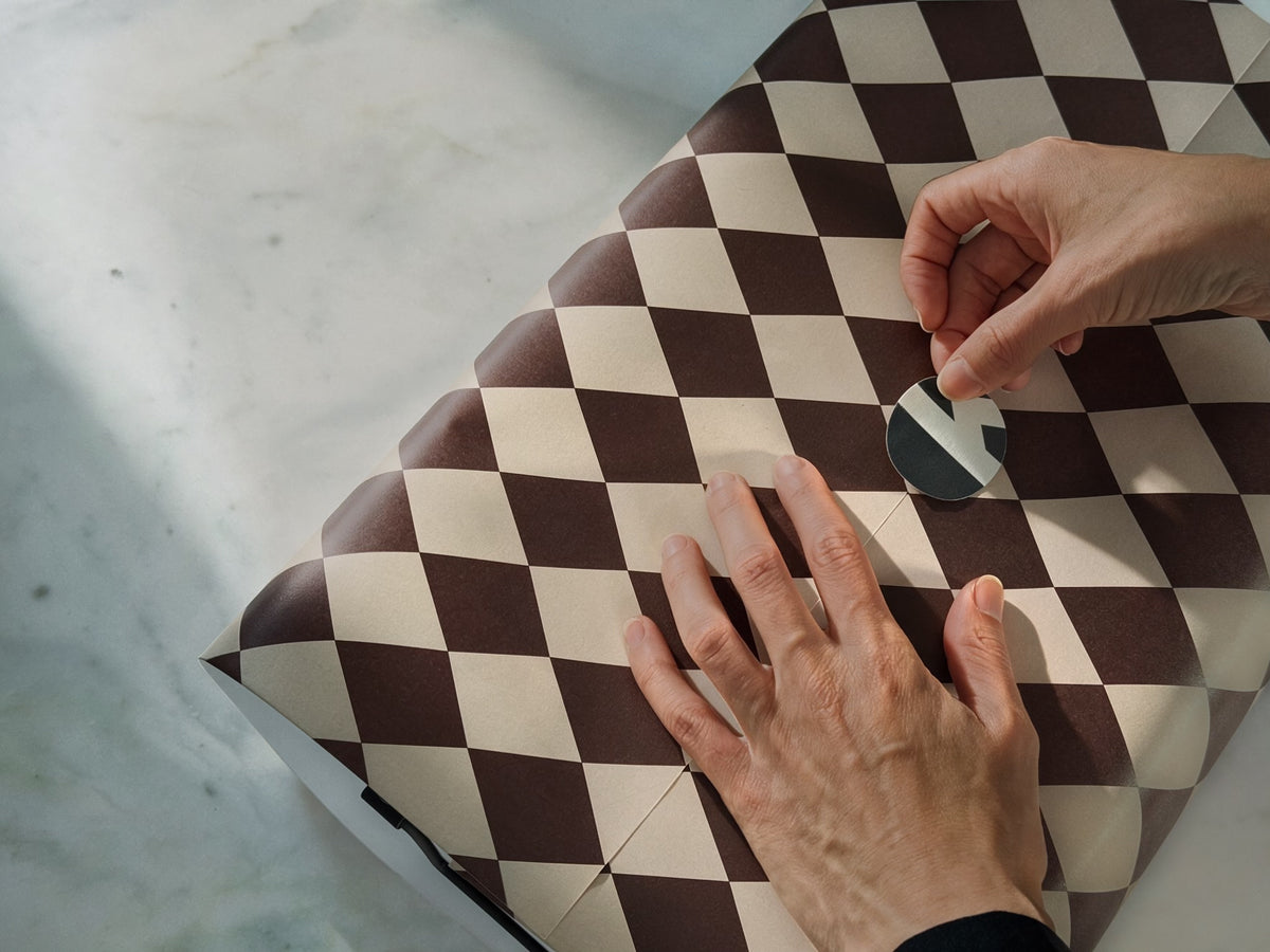A close-up of a person's hands applying a small black and white circular sticker to a dark red and taupe checkered patterned surface, likely a gift box.