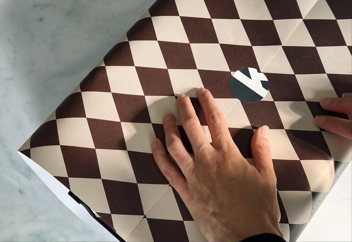 A close-up of a laptop wrapped in a dark red and taupe checkered patterned wrap, with a hand resting on top of it against a light marble surface.