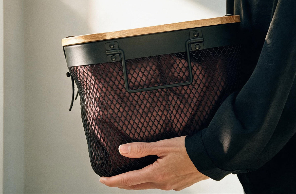 A person in a black long-sleeve top holds a black mesh storage basket with a light wood top and black metal hardware against a softly lit neutral background.