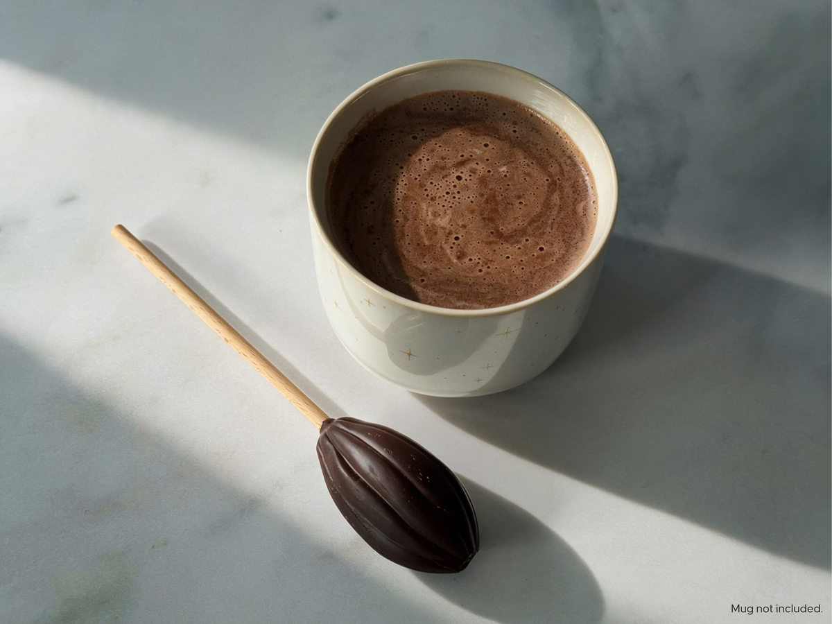 A white ceramic mug filled with a frothy hot chocolate drink sits on a light marble surface beside an oval chocolate-ended stick with a wooden handle.
