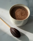 A white ceramic mug filled with a frothy hot chocolate drink sits on a light marble surface beside an oval chocolate-ended stick with a wooden handle.