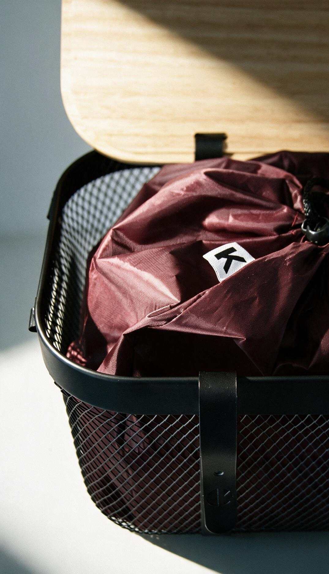 A black wire storage basket with a wooden lid, containing a maroon drawstring bag with a small white square label featuring a stylized 'K' logo, photographed in dramatic sunlight on a white surface.