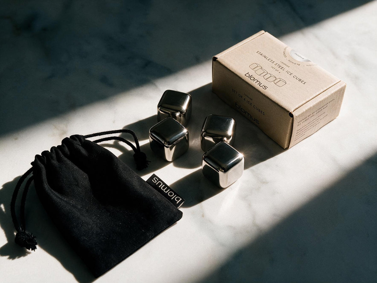 Four metallic ice cubes, a black drawstring pouch, and a kraft paper box on a marble surface in dramatic sunlight and shadow. The pouch and box are branded Blomus.