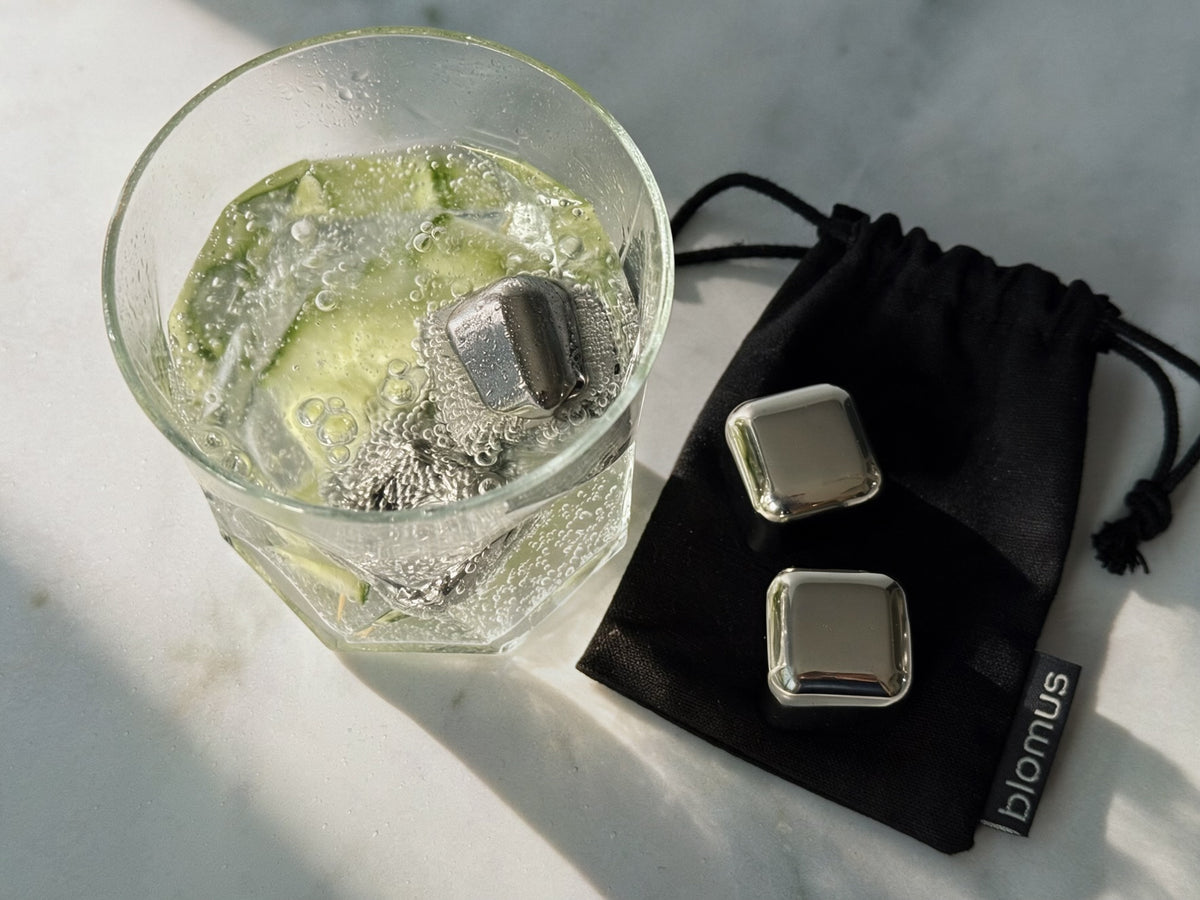 Two silver metal ice cubes shown next to a glass of cocktail with more metal ice cubes, with a black drawstring pouch branded 'blomus' on a light marble surface.