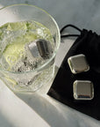 Two silver metal ice cubes shown next to a glass of cocktail with more metal ice cubes, with a black drawstring pouch branded 'blomus' on a light marble surface.