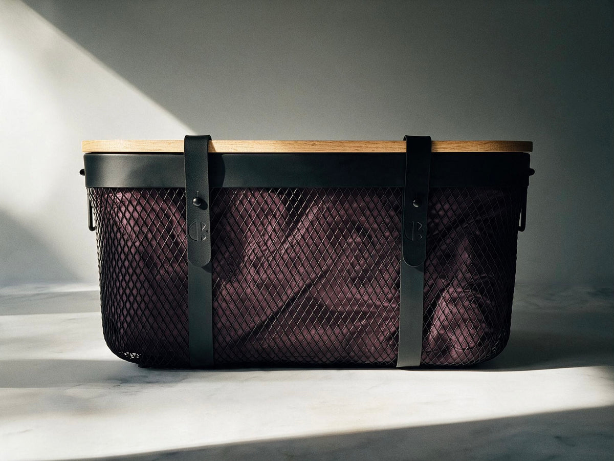 A structured mesh storage basket with a light wood top, black leather straps, and a dark burgundy mesh body, photographed in dramatic side lighting on a gray background.
