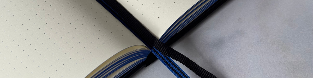 An open dotted journal with cream-colored pages, a black elastic closure, and a blue-and-black striped ribbon bookmark, photographed on a light surface with strong natural shadows.