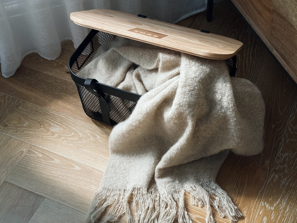 A black wire storage basket with a light wood lid sits on a wood floor next to a white curtain, with a beige knit throw blanket draped out of the basket.