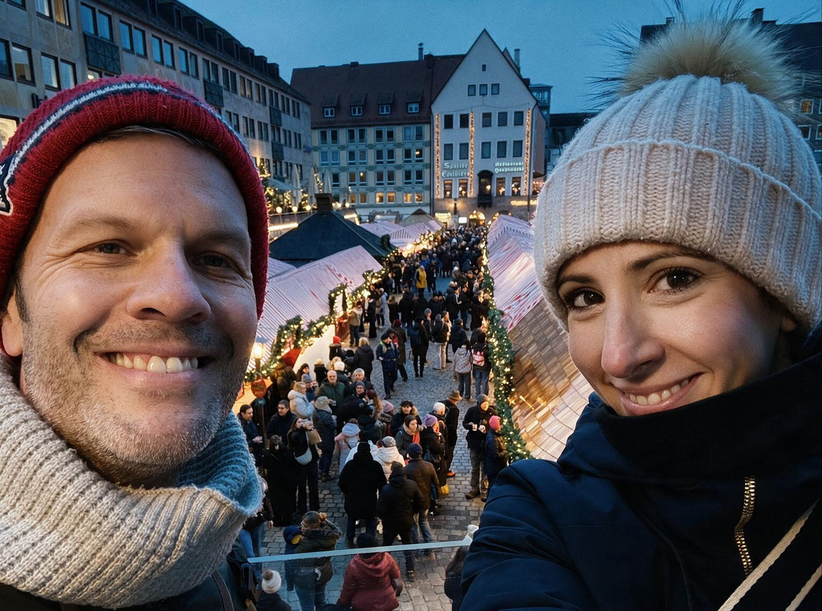 The founders taking a selfie above Nuremberg Christmas Market at dusk, with the man wearing a red knit beanie and light gray knit neck warmer, and the woman wearing a cream knit beanie with a pom-pom and a dark navy jacket.