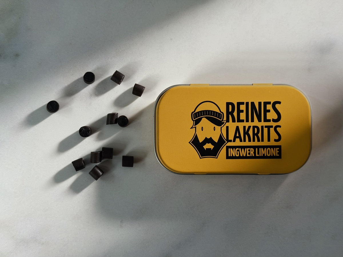 A yellow metal tin of Lemon and Ginger Liquorice sits on a light marble surface with several dark liquorice pearls scattered to the left. The tin features a black graphic of a bearded man wearing a beanie and the text 'REINES LAKRITS INGWER LIMONE'.