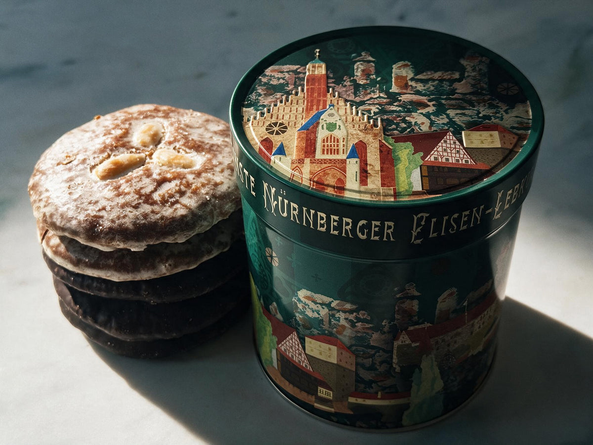 A stack of round gingerbread sits beside a decorated cylindrical tin labeled 'Nürnberger Elisen-Lebkuchen', photographed on a light marble surface with strong sunlight and shadows.