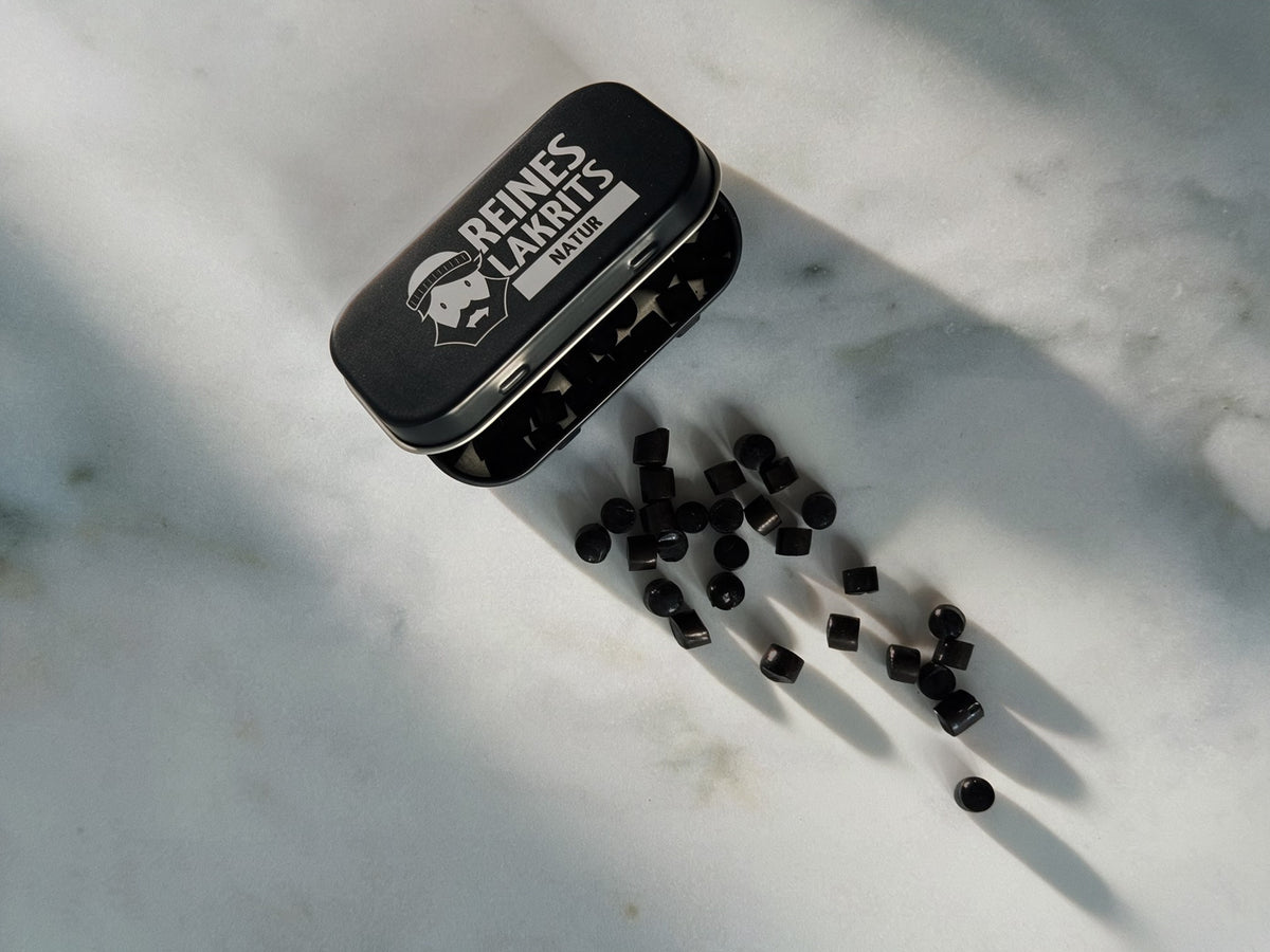 A small black metal tin labeled 'REINES LAKRITS' with a white logo, shown on a light marble surface with several black round liquorice beads spilled out beside it.