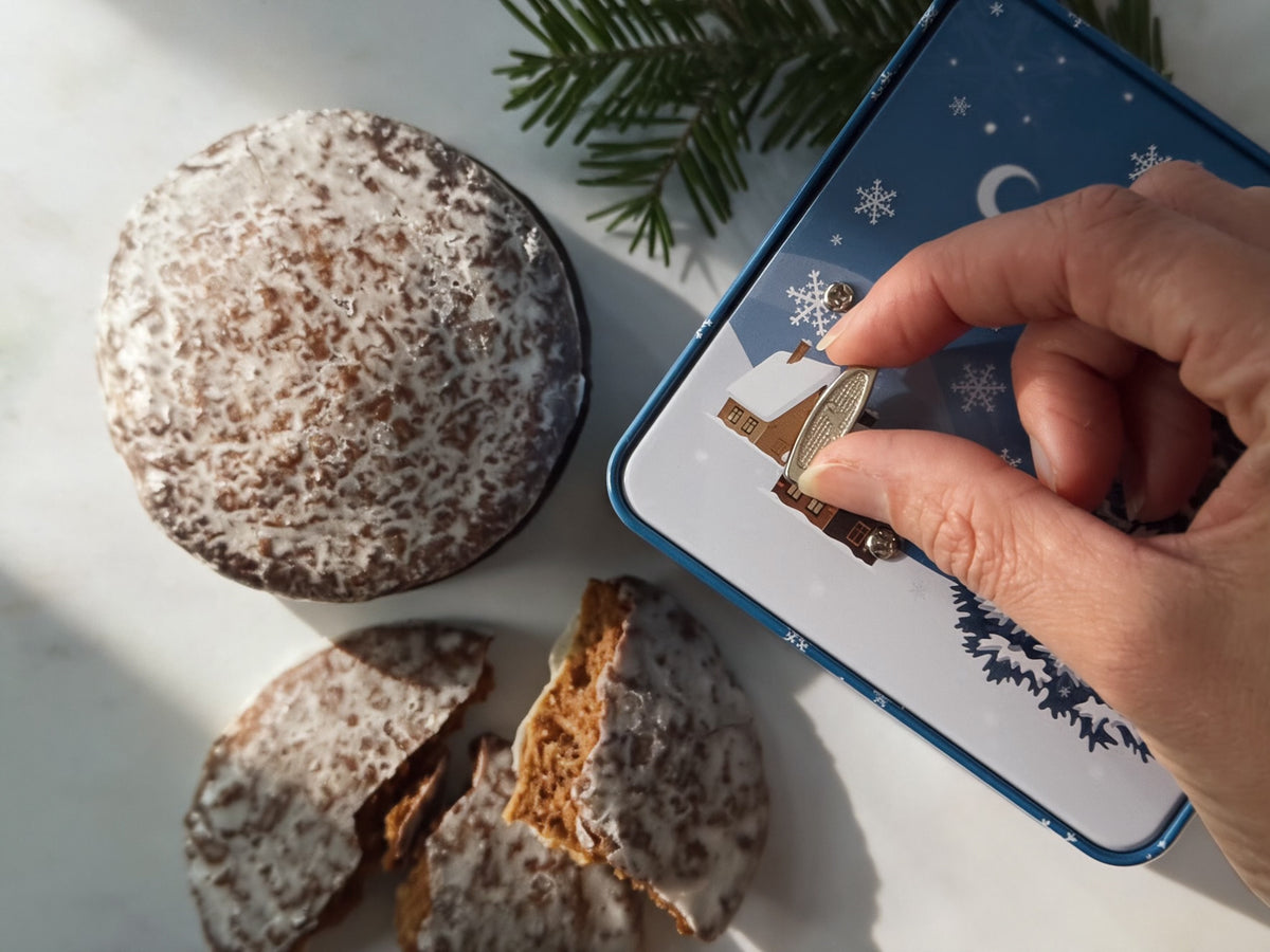 A hand is turning the silver dial of a blue music box-tin with a Christmas tree design. On a white surface next to the tin are decorated Christmas cookies, a stack whole and one broken to show the interior.