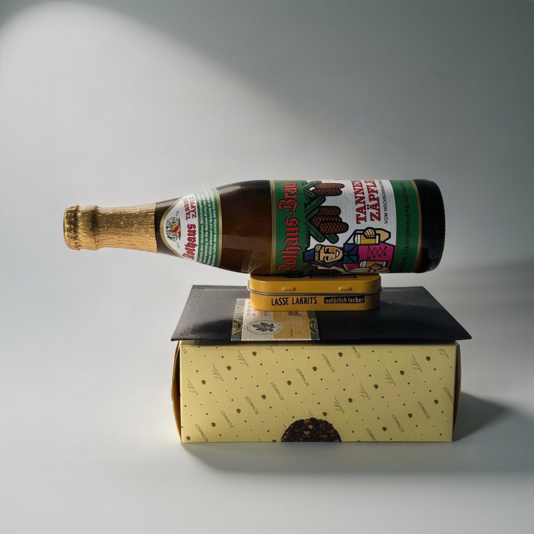 A glass bottle of Rothaus Tannenzaepfle beer with a gold foil neck and colourful label, balanced on top of a yellow Lasse Lakrits tin, which sits on a black rectangular block and a patterned yellow box against a light, plain background.