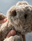 A close-up of a plush Steiff teddy bear being held by two little hands. The bear has shaggy light brown fur, a brown stitched nose, and a yellow Steiff brand tag on its ear.