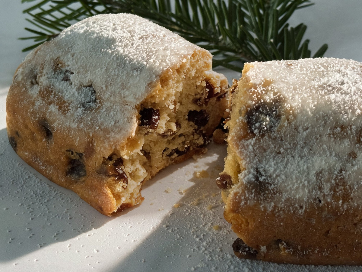 A close-up of a sliced Christmas Stollen, dusted with powdered sugar and showing dried fruit pieces inside, with pine branches in the background.