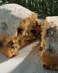 A close-up of a sliced Christmas Stollen, dusted with powdered sugar and showing dried fruit pieces inside, with pine branches in the background.